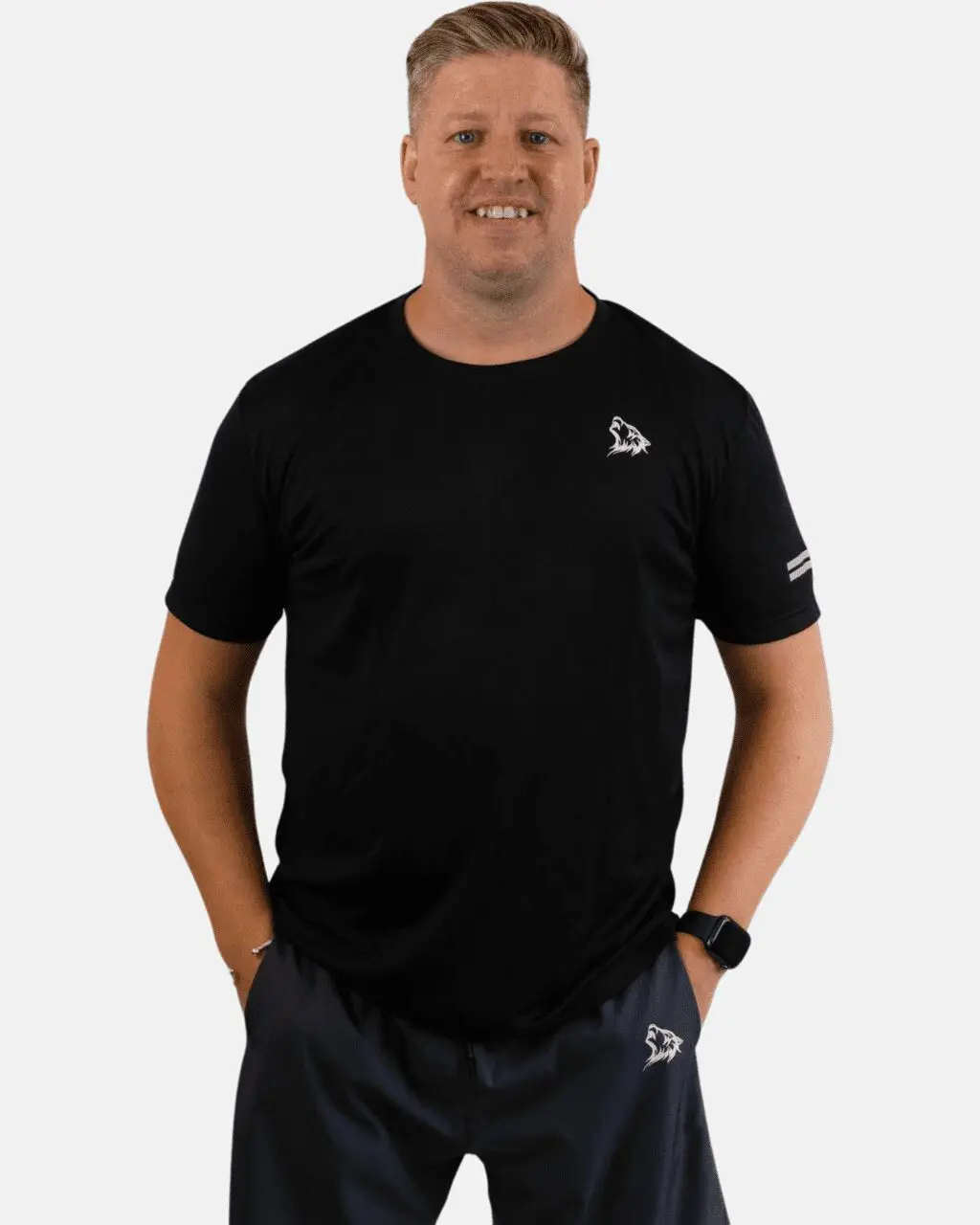 A man wearing black shirt and shorts standing with hands in his pockets.